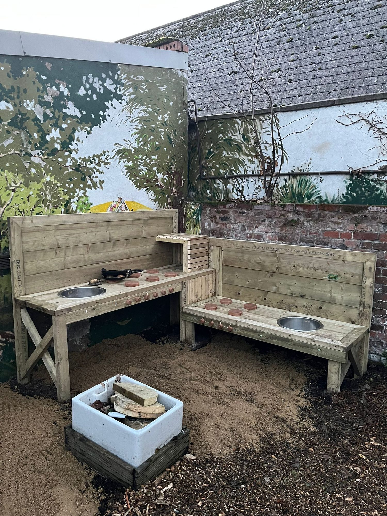 Handmade timber mud kitchen with bowls and play pots against a painted woodland mural