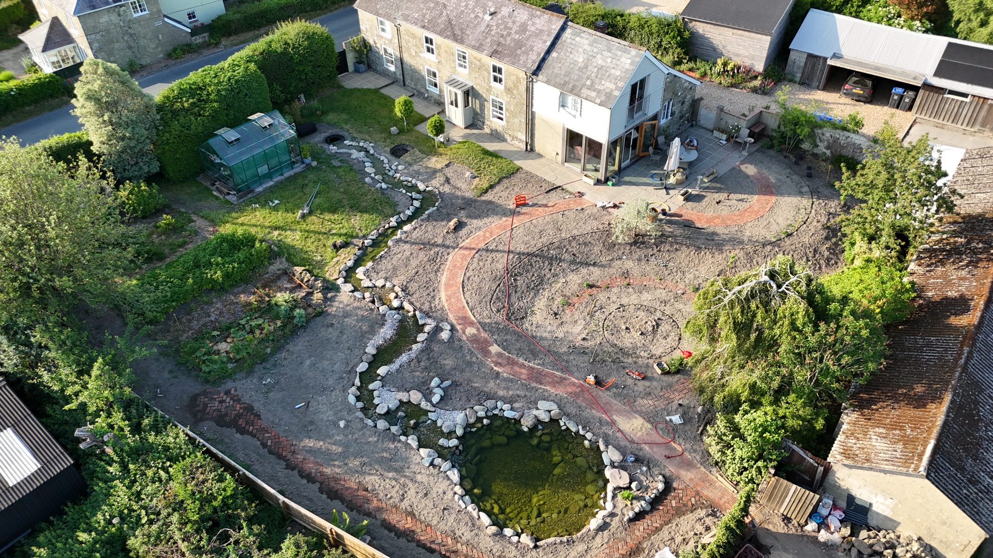 Aerial view of a country-house garden transformation in progress, with a newly dug wildlife pond and a winding clay-paver path