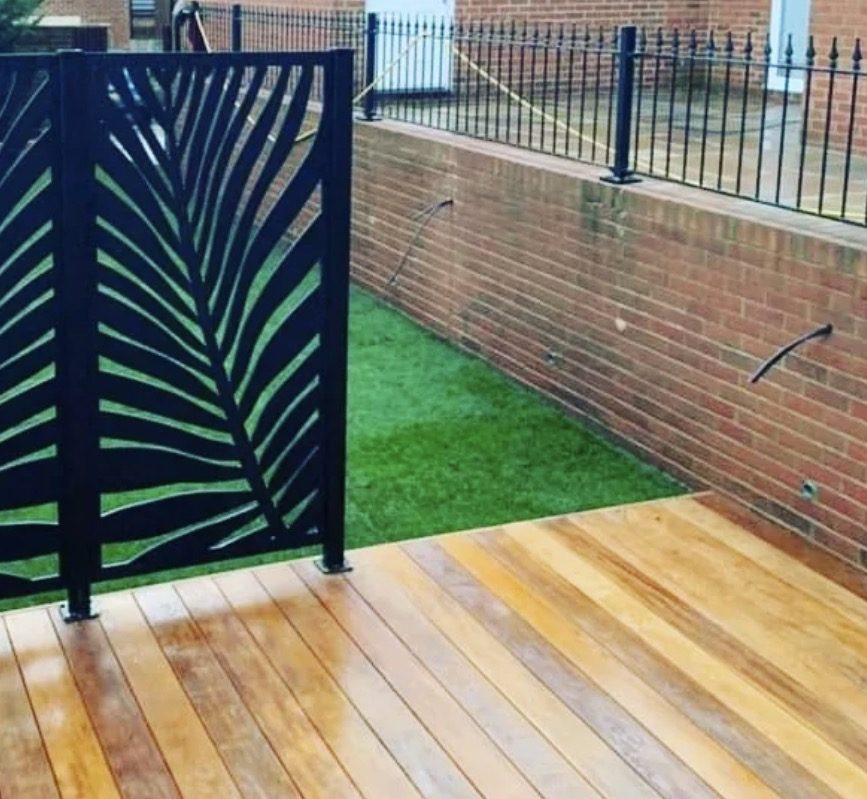 Hardwood decking with a black laser-cut palm-leaf privacy screen and artificial lawn against a brick retaining wall