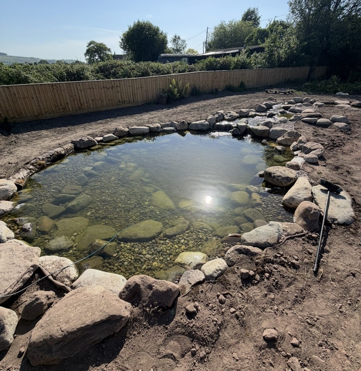 Natural wildlife pond with clear water over a pebble base, boulder edging and a timber fence in rural Wiltshire