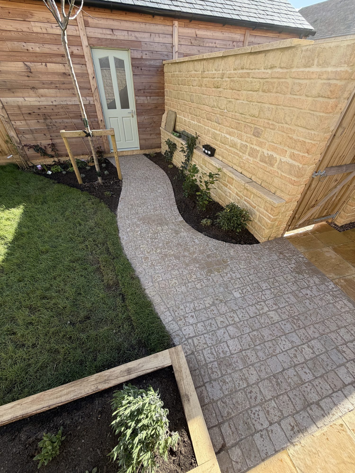 Curved tumbled cobble-sett path leading to a Cotswold stone cottage with an oak sleeper raised bed and new lawn